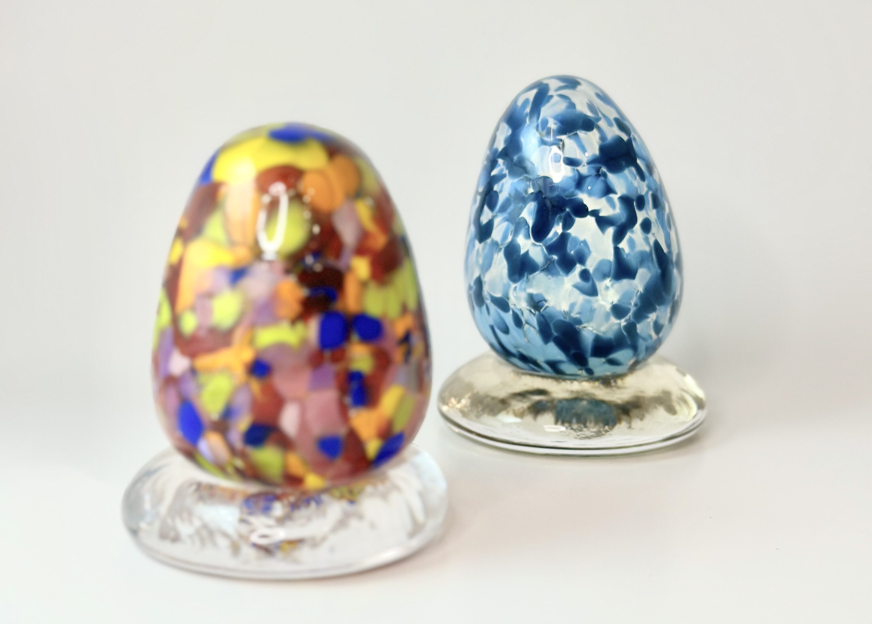 Two blown glass eggs, one in blues, one in bright festive colors, both with a clear glass foot