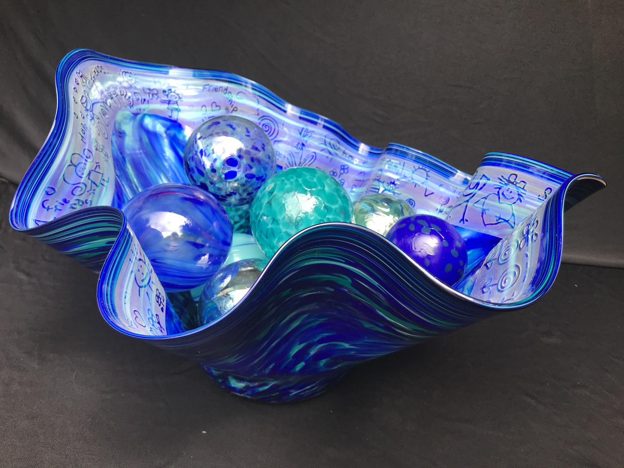 Large fluted blown glass bowl with ornaments nestled in it. The bowl has engravings of children's adrawings of home around the edge.